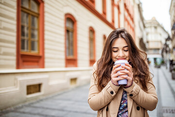 Lifestyle portrait of a young stylish woman in red shirt with coffee cup and smart phone walking on the street in the old city. Young stylish woman drinking coffee to go in a city street