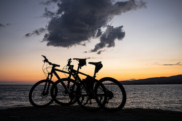 Fototapeta na wymiar Two mountain bikes stand on the edge of a cliff, where you can see a bright orange sea sunset, mountains and large clouds. Recreation and sports in the resort city of Gelendzhik