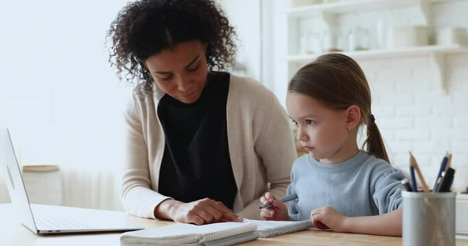 Smiling young biracial foster mother or babysitter checking homework of smart little primary school learner. Focused african american teacher educating small cute girl at private lesson at home.