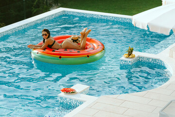 sexy woman relax in swimming pool outdoors at hot summer day. Sexy female in swimsuit on vacations lay on inflatable mattress in swimming pool outdoors at sunset with pool toys. Summer vacation time. 