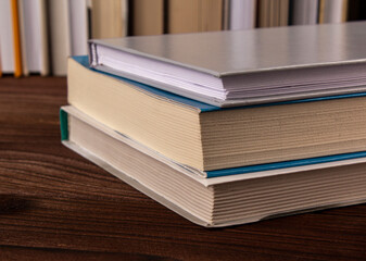 High angle close up of stack of books pile on wooden surface against the background of books standing in a row. Selective focus. Books stacked on a dark background. Novels, tales.