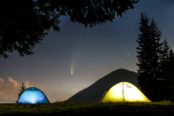 Two brightly lit tourist tents on forest clearing in mountains with starry sky and Neowise comet with light tail.