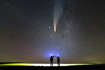 Small silhouettes of two scientists with flashlight on heads pointing bright beam of light on starry sky with C/2020 F3 (NEOWISE) comet with light tail. Space exploration concept. - obrazy, fototapety, plakaty