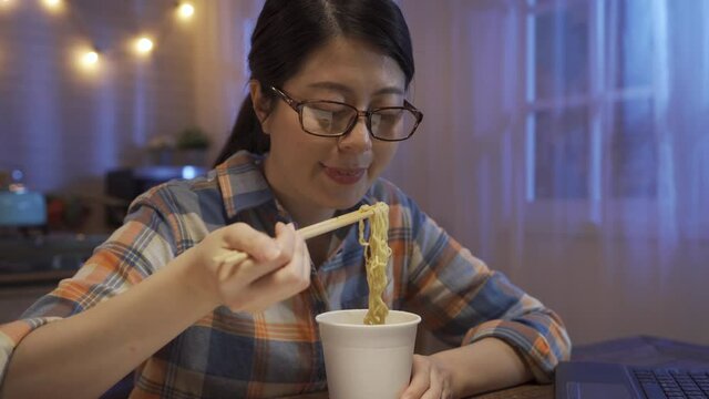 Young female worker sitting at laptop computer eats instant ramen noodles late in evening. Woman in glasses studying online overtime at night fast dinner as bedtime snack. concept unhealthy junk food