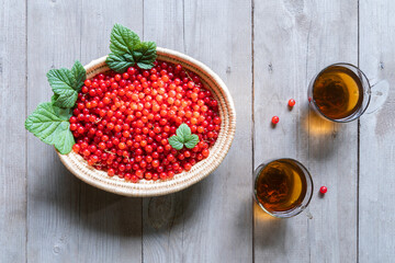 Wicker basket filled by plenty of red currant with leaves on a sun light flecks and herbal tea cups on wooden rustic table background