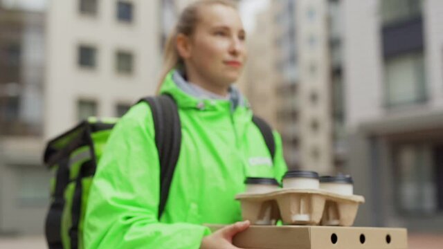 Tilt up tracking slow motion shot of female food courier in green uniform carrying pizza boxes and takeout coffee cups in her hands while walking down street delivering order, thermal bag behind back