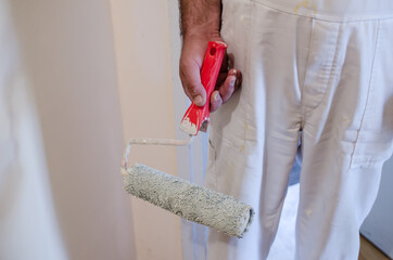 hand of working man in white, standing in empty room, holding paint roller . Painting apartment, renovating with white color paint