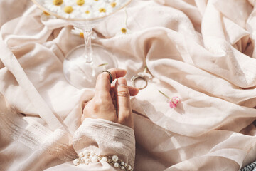 Hand with jewelry on background of soft beige fabric with stylish wineglass with daisy flowers in...