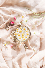 Tender floral aesthetic. Daisy flowers in water in glass cup on background of soft beige fabric with wildflowers and jewelry.  Creative summer image with space for text. Bohemian mood
