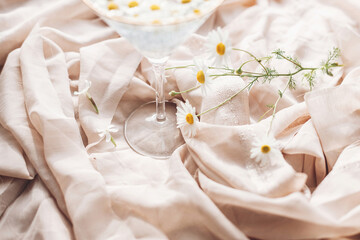 Daisy flowers and white wildflowers at wineglass stem on background of soft beige fabric. Tender floral aesthetic. Creative summer image. Bohemian mood