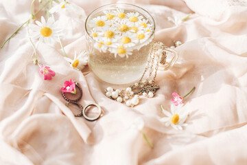 Daisy flowers in water in glass cup on background of soft beige fabric with wildflowers and jewelry. Tender floral aesthetic. Creative summer image with space for text. Bohemian mood