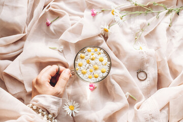 Hand with jewelry holding glass cup with daisy flowers in water on background of soft beige fabric with wildflowers. Tender floral aesthetic. Creative summer image, flat lay. Bohemian mood
