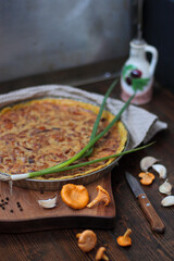 Mushroom quiche in a metal form on a wooden cutting board. Small knife. Green onion on the pie. Olive oil in ceramic bottle. Some chanterelle mushrooms and garlic on the table.