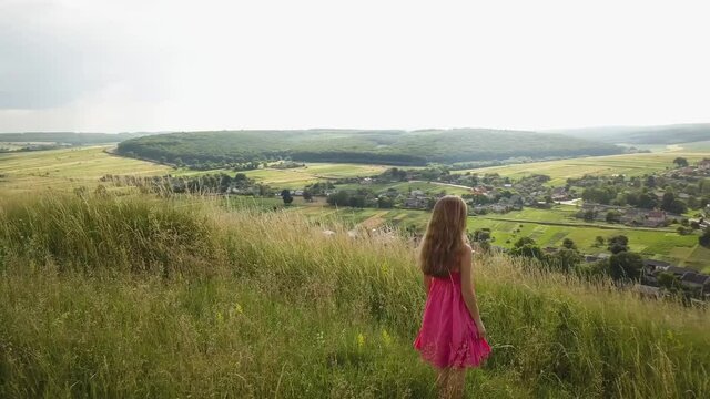 Yong woman with long hair in red dress walking in summer field with tall green grass on rural hill environment.