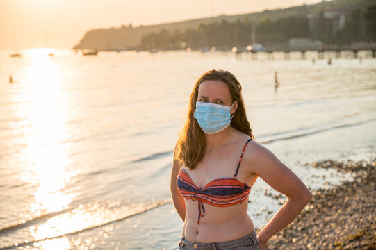 Young woman wearing medical mask at the beach during sunset.