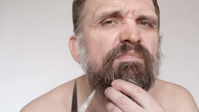 Mature man with beard on fave tries to cut it with scissors. Everyday care of face.