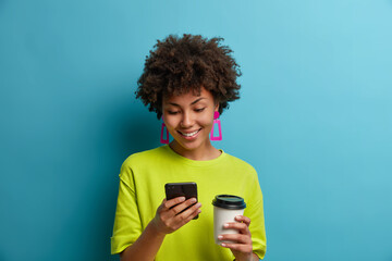 Happy positive ethnic young woman types message or dials telephone number on phone, drinks takeaway...