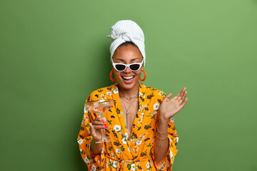 Joyful positive housewife wears wrapped towel on head and yellow dress with flower print, drinks...