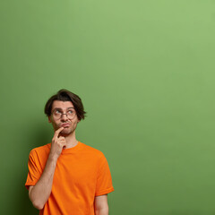 Fototapeta na wymiar Pensive indecisive adult man looks upwards and keeps finger near mouth, dressed in casual orange t shirt, poses against green background with copy space for your promotion or advertising content