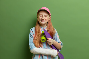 Children, dangerous hobby and injuries concept. Pleased redhead girl closes eyes and feels happy, holds skateboard under arm, got trauma during skateboarding, has active summer rest, isolated on green © WHstudio Leushin N