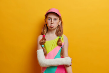 Sad gloomy freckled girl wears colorful bathing suit and cap, has broken arm in cast, spoiled...