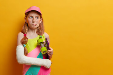 Puzzled redhead girl holds longboard under arm, poses with skate and broken hand in cast, has unlucky day and looks displeased aside, isolated on yellow background, got injured after doing trick © wayhome.studio
