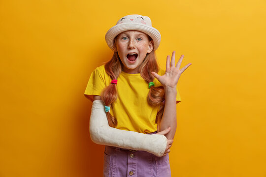 Portrait of joyous redhead girl waves palm in hello gesture, says hi to parents, being in good mood, wears summer outfit, cast on broken arm after falling during rollerblading, isolated on yellow wall