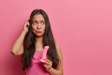 Indecisive Asian woman rubs head chooses dildo in sex shop holds silicone rosy phallic object to provide sexual pleasure via penetration finds perfect size for herself, subsititues real mens genitalia
