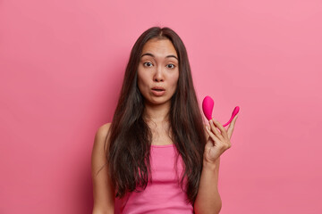 Astonished brunette female holds modern remote app controlled smart vibrator, visits sex shop and buys necessary accessories to satisfy herself, dressed casually, poses against pink background