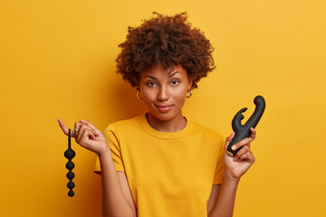 Pretty young woman gets sexual experience with sex toy accessories, holds anal beads to stimulate erogenous zone in your anus, vibrator to reach orgasm, stands against vivid yellow background