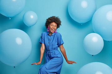 Carefree joyful curly woman dances happily, dressed in blue dress, chills at party around inflated helium balloons, feels playful, enjoys favorite holiday, has upbeat festive mood. Moment of joy - obrazy, fototapety, plakaty