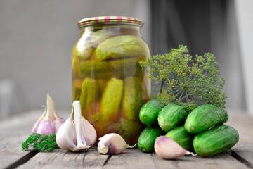 A jar of canned cucumbers on a wooden table, next to fresh cucumbers, garlic and herbs. The concept of harvesting vegetables for the winter.