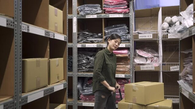 young girl worker in uniform moving and taking out cardboard box and lifting on shelves in warehouse. asian korean female employee search empty place and putting parcel from trolley in stockroom.