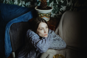 Young sad girl with a gray knitted sweater sits in a room on a chair with wraps