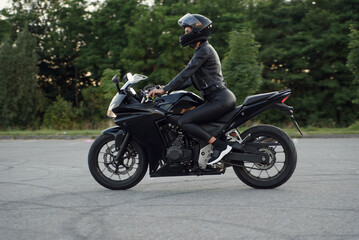 Stylish woman in black leather jacket, pants and full-face protective helmet rides on sports motorcycle at urban outdoors parking.