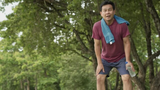 Asian old age man stop and resting during running at the park, standing under trees and rest after exercising, body wellness tired exhausted, men heath, elderly retired life, afternoon cardio workout