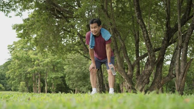 Asian old age man stop and resting during running at the park, standing under trees and rest after exercising, body wellness tired exhausted, men heath, elderly retired life, afternoon cardio workout