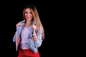 A young smiling girl in a denim jacket and red trousers isolated on a black background