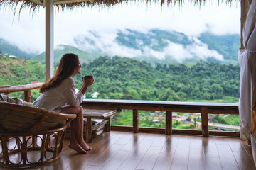 Portrait image of a beautiful asian woman holding and drinking hot coffee , sitting on balcony and looking at mountains and green nature