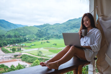 A beautiful asian woman working and typing on laptop computer while sitting on balcony with mountains and green nature background