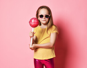 Frolic 6-7 y.o. kid girl in yellow t-shirt, shiny pink leggings and sunglasses holds big heavy pink lollipop