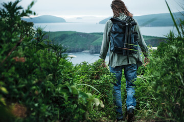 back view of a young long-haired man with a backpack and a photo tripod walking along a trail through the grass at the edge of a cliff with sky and sea background.. Travel and outdoor concept.