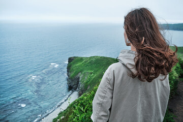 back view of a girl standing in the grass at the edge of a cliff with sky and sea background. Journey and outdoor concept.