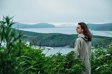 back view of a girl standing in the grass at the edge of a cliff with sky and sea background. Journey and outdoor concept.