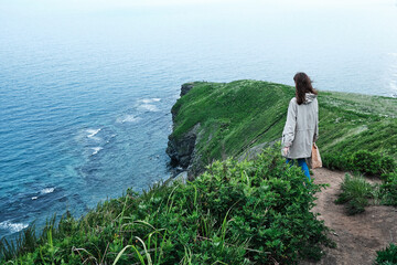 back view of a girl walking along a trail through the grass at the edge of a cliff with sky and sea background. Travel and vacation concept.