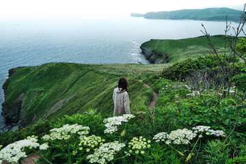 back view of a girl walking along a trail through the grass at the edge of a cliff with sky and sea background. Travel and vacation concept.