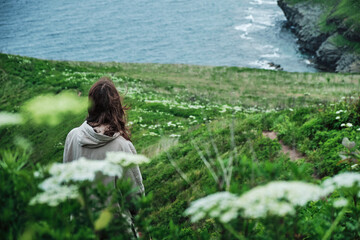 back view of a girl standing in the grass at the edge of a cliff with sky and sea background. Journey and outdoor concept.
