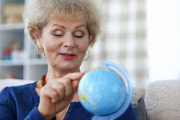 Elderly woman hold blue globe in her hand and point to continent with her index finger. Traveling around the world at retirement age. Exploration of world map. Travel agency carries out selection of