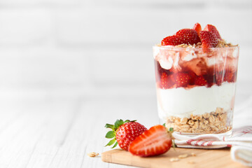 Homemade layered dessert with fresh strawberries, cream cheese or yogurt, granola and strawberry jam in glass on white wood background. Healthy organic breakfast or snack concept. Selective focus.