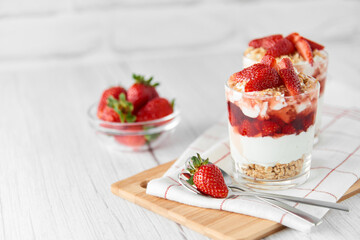 Homemade layered dessert with fresh strawberries, cream cheese or yogurt, granola and strawberry jam in glasses on white wood background. Healthy organic breakfast or snack concept. Selective focus.